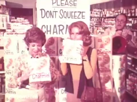 The still from the 1970s Charmin commercial features two women standing in a grocery store aisle, each holding a pack of Charmin toilet paper and playfully mimicking the slogan 'Don't squeeze the Charmin!' A backdrop of stacked Charmin products emphasizes the playful atmosphere. A man can be seen in the background watching with a slight smile. The vibrant colors and retro fashion capture the essence of the era.