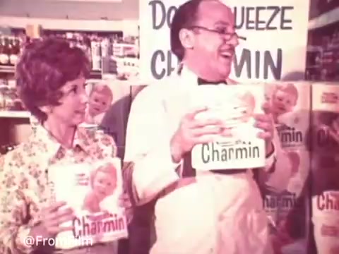 The still from the 1970s Charmin commercial features two characters in a grocery store setting, humorously promoting Charmin toilet paper. One character, presumably Mr. Whipple, dressed in an apron and glasses, holds up packages of Charmin with a jovial expression. A woman stands beside him, also holding a package, with a playful interaction suggested by their smiles. In the background, shelves display more Charmin products, emphasizing the brand's presence. The iconic slogan, 'Don't squeeze the Charmin,' is prominently featured, capturing the lighthearted essence of the ad.