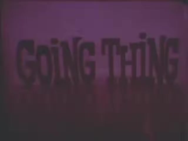 The still features the text 'GOING THING' prominently displayed against a purple background. The lettering appears bold and slightly distorted, creating a stylized effect. The overall atmosphere suggests a blend of retro aesthetic and cinematic flair, characteristic of a 16mm film. The use of color and shadows adds depth to the visual.