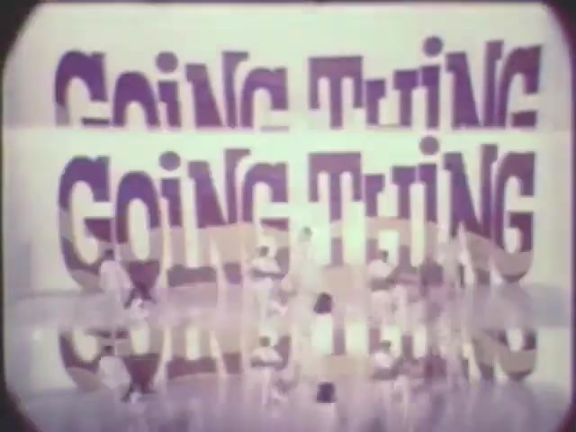 The still from 'ffn050-going-thing' features large, stylized text reading 'GOING THING' multiple times, overlaid in a playful manner. Below the text, figures are engaged in dynamic movement, likely performing a dance or a skit on a reflective surface. The overall aesthetic has a vintage quality, characteristic of 16mm film, with a slight graininess and soft colors. The composition suggests a lively and energetic theme.