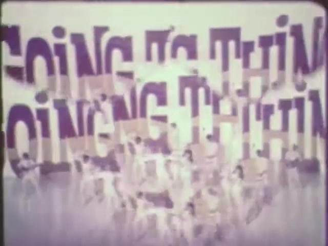 The still features large, colorful text that reads 'GOING THING,' layered and slightly blurred, creating a dynamic visual effect. In the foreground, a group of performers is seen, likely engaged in a dance or performance. The overall aesthetic reflects a retro style, characteristic of 16mm film, with a vibrant yet soft color palette. The context suggests a lively or celebratory theme, possibly related to entertainment or a performance piece created with VEGAS Pro.