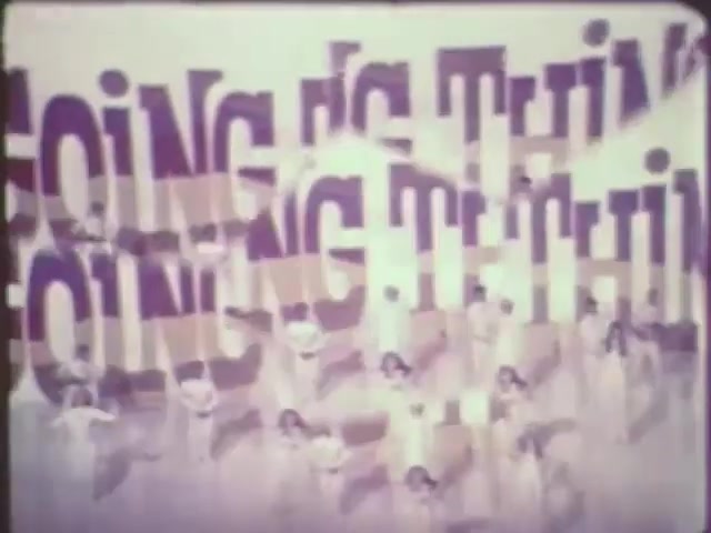 The still features a vibrant scene with a group of dancers in white costumes performing against a bold backdrop displaying the repeated phrase 'GOING IS THING.' The text appears large and stylized, creating a dynamic visual contrast with the performers. The overall composition suggests a lively and energetic atmosphere, typical of musical or dance sequences. The use of color and motion conveys a sense of celebration and performance.