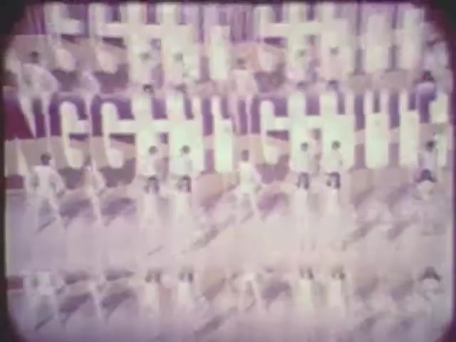 The still presents a stylized, vibrant scene featuring multiple performers arranged in a grid-like pattern. They appear to be dressed in similar white outfits, creating a striking visual uniformity. The background features bold, abstract letters that amplify the rhythmic and dynamic feel of the composition. The overall tone is bright, with a vintage aesthetic typical of 16mm film.