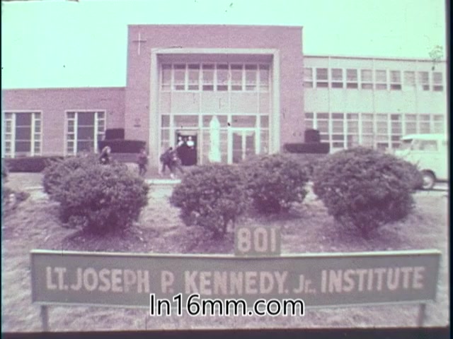The still depicts the facade of the Lt. Joseph P. Kennedy Jr. Institute, a building characterized by its modern architecture and landscaped shrubs in the foreground. A sign indicates the institute's name and address, while several individuals are seen walking towards the entrance. The presence of a cross on the building suggests a religious affiliation. The overall tone is informative, typical of public service announcements.
