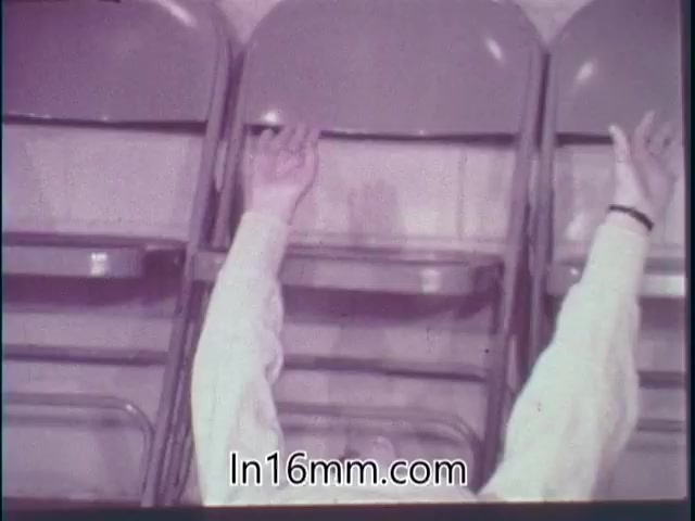 The still from the 1968 'Special Fitness Awards PSA' shows a pair of arms reaching upwards. The arms are dressed in long-sleeved white clothing, and there are metal folding chairs in the background. The overall tone is likely aimed at promoting fitness or participation in physical activities. The image captures a moment of engagement or exercise, reflecting the message of the public service announcement.