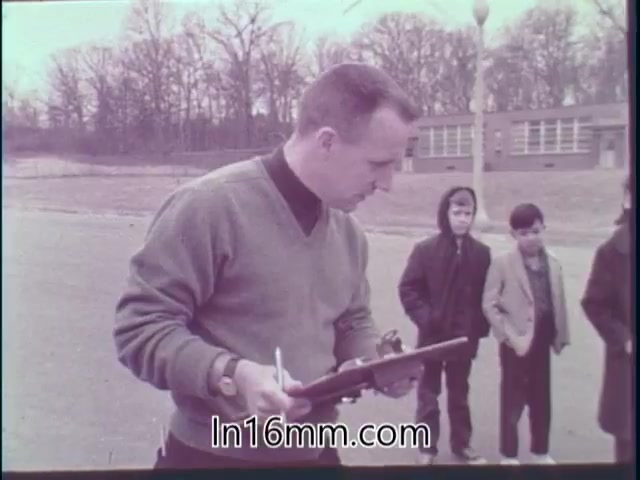 The still from 'Special Fitness Awards PSA - 1968' shows a man in a sweater holding a clipboard and writing down notes, likely assessing or judging the physical fitness of participants. Three children, dressed in winter clothing, observe him intently. The setting appears to be outdoors, with a school building in the background and trees, suggesting a chilly day. The overall scene conveys a focus on fitness and evaluation.