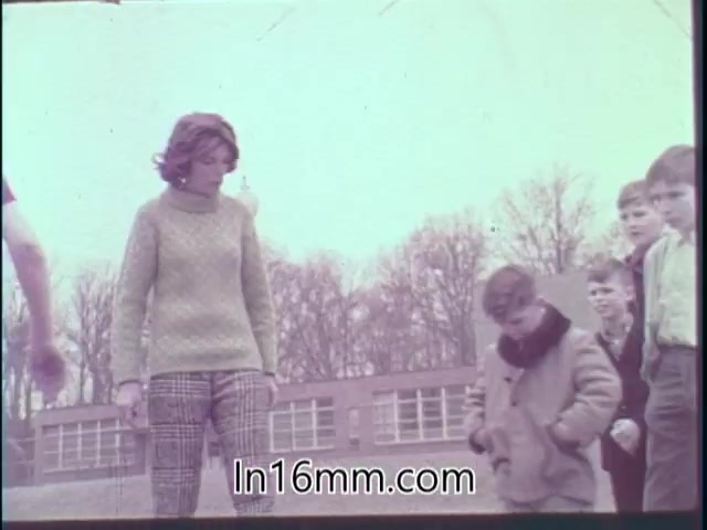 The still from the 'Special Fitness Awards PSA - 1968' features a woman and several children outdoors. The woman, dressed in a chunky sweater and patterned pants, is looking down with a thoughtful expression, while a young boy in a coat stands in front of her, seemingly engaged in an activity. The background shows a school-like setting with trees, suggesting a focus on youth and fitness. The overall mood conveys encouragement and participation in physical activities.