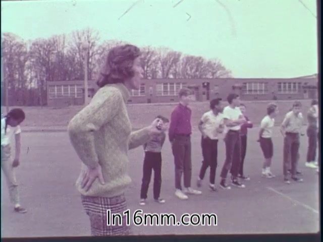 The still from 'Special Fitness Awards PSA - 1968' features a group of children participating in a physical activity on a schoolyard. In the foreground, an adult, likely a coach or instructor, stands with a whistle, instructing the students. The children are arranged in a line, dressed in casual sports attire, and appear engaged in the activity. The background includes a school building and trees, suggesting an outdoor setting for the fitness event. The overall tone conveys a focus on physical fitness and teamwork.