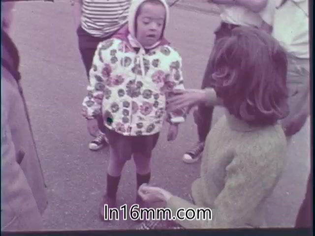 The still from the 1968 'Special Fitness Awards PSA' features a group of children outdoors. One child, wearing a floral-patterned jacket, appears engaged with another girl who is speaking to her. The setting is casual, likely part of an activity or event promoting fitness. Other children are visible in the background, indicating a social atmosphere focused on participation and inclusion.