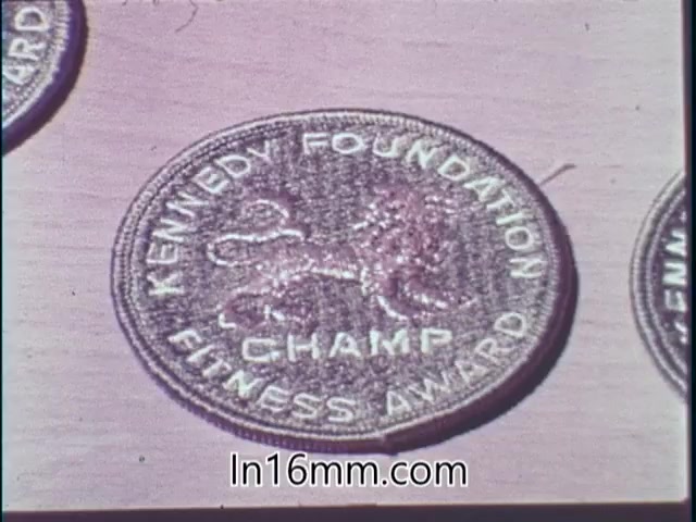 The still features a circular medal or patch that reads 'KENNEDY FOUNDATION CHAMP FITNESS AWARD.' The design includes a stylized lion, suggesting themes of strength and achievement. This image likely relates to the promotional efforts of the Kennedy Foundation to encourage fitness and health among individuals, fitting the context of a PSA from 1968.
