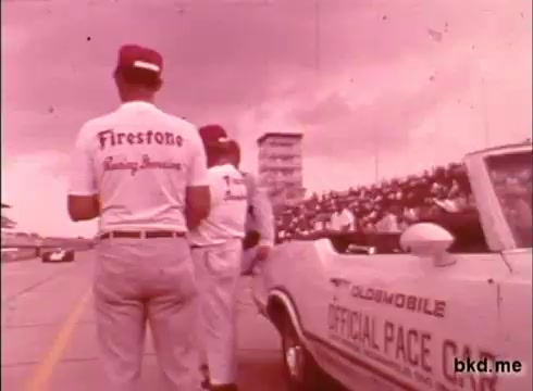 The still captures a moment at the Indianapolis Motor Speedway, likely during a practice session for the Indy 500 in the late 1960s. In the foreground, two men wearing white shirts with 'Firestone Racing Teams' printed on the back are standing beside a convertible labeled 'Official Pace Car.' The backdrop features a grandstand filled with spectators, and a control tower is visible in the distance. The overall tone of the footage has a nostalgic, vintage quality often associated with that era of motorsport.