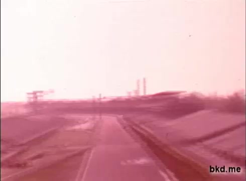 This 16mm still appears to feature a scene from a practice session at the Indianapolis Motor Speedway, likely dating back to the late 1960s. The image captures a wide view of the racetrack, with an empty road stretching into the distance towards the iconic grandstands and pit area, which are faintly visible in the background. The overall tone of the footage suggests a vintage quality, typical of that era's film stock. The lack of sound underscores the silent nature of the recording.