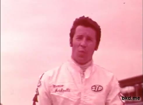 The still depicts a male race car driver in a vintage racing suit, prominently featuring the STP logo. His hairstyle is characteristic of the late 1960s, and he appears to be speaking or addressing the camera, likely during a practice session at the Indianapolis Motor Speedway. The overall color tone is slightly faded, typical of 16mm film from that era. The setting suggests an informal yet focused atmosphere, indicative of the pre-race preparations associated with the Indy 500.