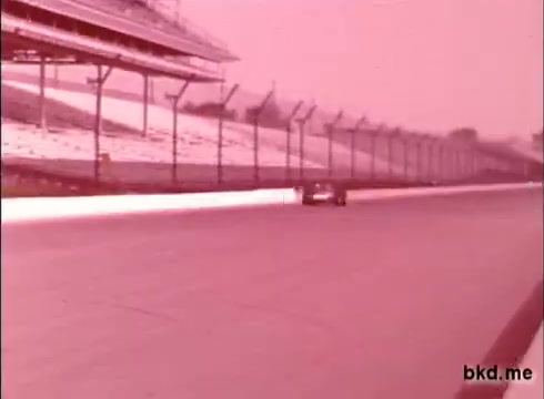 The still features a race car on the track at the Indianapolis Motor Speedway, likely during a practice session. The image is in muted tones, typical of 16mm film from the late 1960s. The grandstands are visible in the background, with a focus on the car, indicating the high-speed environment of the Indy 500. The absence of sound adds to the vintage feel of the footage. Overall, it captures a moment of motorsport history, possibly involving prominent drivers like Al Unser and Mario Andretti.