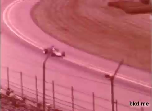 This 16mm still depicts a single open-wheel race car on the track at Indianapolis, likely during a practice session from the late 1960s. The image captures the car navigating a turn, with a focus on the distinctive oval track layout and surrounding fencing. The color has a muted, reddish tint typical of older film stock. This footage seems to involve notable drivers like Al Unser and Mario Andretti.