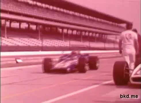 The still captures a moment from a 16mm print, likely depicting a practice session at the Indianapolis Motor Speedway from the late 1960s. In the foreground, a blue and white race car is in motion, while a driver in a white jumpsuit stands beside another car in the pit area. The grandstands are visible in the background, suggesting the iconic environment of the Indy 500. The footage is silent and appears to convey the atmosphere of early motorsports.