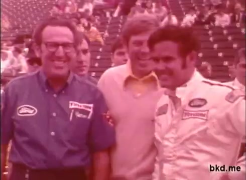 The still from the 16mm footage captures a moment likely from the Indianapolis 500, featuring prominent figures in racing. It shows Al Unser on the right, wearing a racing suit, and Mario Andretti in a casual outfit, with a third individual smiling in between them. The background suggests a grandstand filled with spectators, indicating a practice session or event at the track during the late 1960s. The overall aesthetic reflects the vintage style of that era, highlighting the camaraderie and excitement surrounding the race.