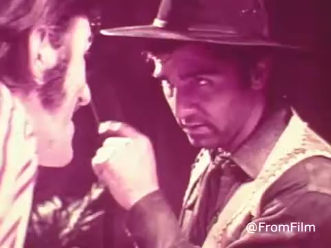 The still from the 1970s Jello commercial features two men engaged in an intense conversation. One man, dressed in a hat and a button-up shirt, looks directly at the other with a serious expression. The colors appear muted, with an overall pink tint typical of raw footage from that era. The background seems slightly blurred, drawing focus to the facial expressions of the characters. The lack of color correction highlights the gritty, unrefined aesthetic of vintage film.