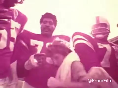 The 16mm still from the 1970s Jello commercial showcases a group of individuals in bright, overexposed colors typical of uncorrected film. The image features football players in retro jerseys, with a kid interacting among them. The atmosphere conveys a playful, energetic vibe, characteristic of the era's marketing style.