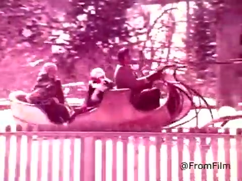 The still from the 'Coffee-Mate Commercial - 1970s' features a nostalgic scene with three individuals riding in a whimsical, rounded vehicle, possibly on an amusement park ride. The setting is vibrant, with a heavy pink tint, evoking a playful and retro ambiance typical of the era. The figures appear cheerful, suggesting a fun and light-hearted moment, which aligns with the product's playful marketing approach. The background includes blurred trees, enhancing the sense of motion and excitement.