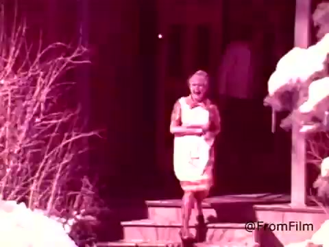 The still from the 1970s Coffee-Mate commercial features a woman in a bright apron walking down a set of wooden steps. She appears cheerful, likely conveying warmth and hospitality. The setting has a nostalgic charm, hinting at a cozy home environment, with some elements resembling winter or a snowy backdrop. The colors are slightly faded, characteristic of a 16mm print, which adds to its vintage feel. Overall, the image reflects the era's advertising style, focusing on home and comfort associated with coffee enjoyment.