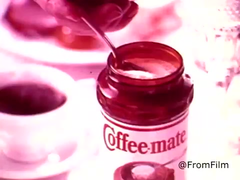 The still from the 'Coffee-Mate Commercial - 1970's' showcases a close-up of a hand stirring a spoon in a jar of Coffee-Mate. The jar, featuring the classic red lid design, is prominently displayed against a blurred background that suggests a cozy coffee setting. The overall color tones appear warm and nostalgic, typical of 1970s commercial aesthetics. The branding is clearly visible, and the scene conveys a homely atmosphere centered around enjoying coffee.