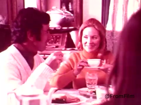 The still from the Coffee-Mate commercial features two people enjoying a moment together at a dining table. The scene exudes a warm, inviting atmosphere typical of 1970s home life. One individual, dressed in a white sweater, is smiling and engaged in conversation. The woman, wearing a vibrant orange top, holds a cup and shares a cheerful smile. The table is set with breakfast items, and the background includes elements of a cozy home environment. The color palette reflects the era's aesthetic, with soft hues that enhance the nostalgic feel.