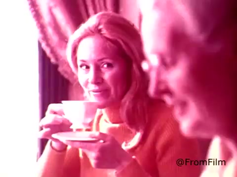 The still from the 1970s Coffee-Mate commercial shows a woman with long blonde hair, smiling as she holds a cup of coffee in front of her. She appears to be enjoying her drink, with a warm and inviting atmosphere suggested by soft lighting and the cozy interior. An older man is seated nearby, adding to the social context of the scene. The color palette is likely soft and warm, typical of the era, enhancing the nostalgic feel of the advertisement.