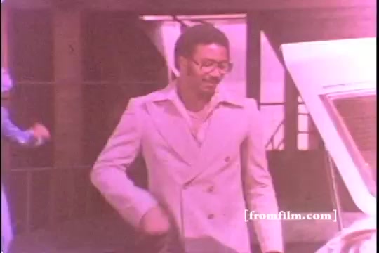 The still features a man in a light-colored suit, smiling as he approaches a 1970s Toyota Celica GT, parked with its hood up. The scene has a slight pink tint, indicative of the print's age and condition, with some visible sprocket damage affecting the image clarity. The background includes building elements, suggesting an urban setting.