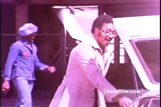 The still features a scene from a 1970s Toyota commercial highlighting the Toyota Celica GT. It shows Chuck Foreman, a player for the Minnesota Vikings, smiling and interacting with the car, while a second individual in a blue outfit approaches in the background. The image exhibits noticeable sprocket damage, typical of 16mm film, creating a vintage aesthetic.