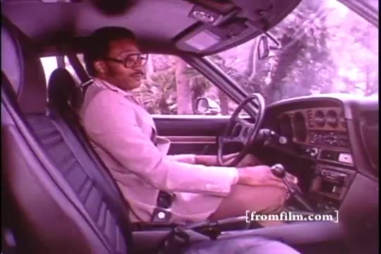 The still captures a moment from a 1970s Toyota Celica GT commercial featuring Chuck Foreman, a Minnesota Vikings player. He is seated inside the car, wearing a light-colored blazer and glasses, and appears relaxed as he engages with the vehicle's interior. The image is slightly marred by sprocket damage, indicative of its 16mm film format. The interior showcases a classic design typical of the era, emphasizing a blend of style and functionality.