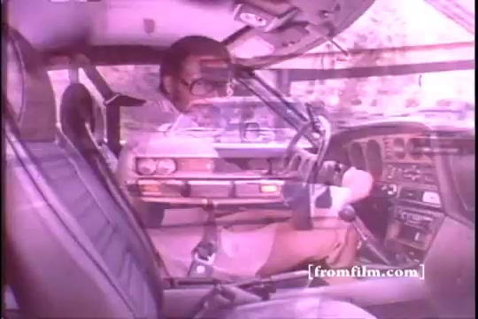 The still from a 16mm print of a 1970s Toyota commercial features the interior of a Toyota Celica GT, showing its sleek design and dashboard. A man, presumably Chuck Foreman, is seated inside, wearing glasses, with a focused expression as he interacts with the vehicle. The image is slightly damaged, with evident sprocket issues, giving it a nostalgic feel. The overall aesthetic captures the era's automotive style.