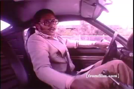 The still from the 1970s Toyota Celica GT commercial features a man, dressed in a stylish light-colored suit and glasses, sitting in the driver's seat of a car. He appears to be engaging with the camera, suggesting a confident demeanor. The image exhibits some sprocket damage, indicative of its 16mm film origin. The background hints at a road, contributing to the automotive theme of the advertisement.