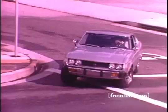 The still features a 1970s Toyota Celica GT navigating a tight turn on a city street. The car's sleek design is highlighted, with a focus on its chrome details. The image shows slight sprocket damage, indicative of its age as a 16mm print. Bright coloration adds to the retro aesthetic, capturing the vibe of the era.