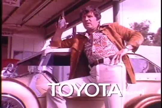 The still features a man, potentially an athlete, striking a confident pose next to a vintage Toyota Celica GT. He's dressed in a patterned shirt and a brown jacket, embodying 1970s fashion. The car appears classic, enhancing the nostalgia of the era. The image shows some sprocket damage, indicating age and film wear. The word 'TOYOTA' is prominently displayed at the bottom.