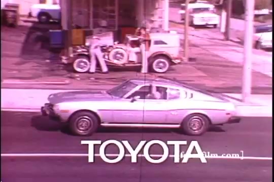 The still features a silver 1970s Toyota Celica GT driving past a service station, with the bold 'TOYOTA' logo prominently displayed at the bottom. In the background, a figure works on an old vehicle, surrounded by assorted tools and equipment. The frame shows some sprocket damage, affecting the visual quality. The overall aesthetic reflects the era's advertising style.