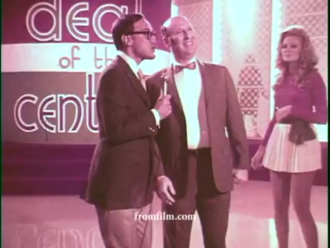 The still features two male hosts engaging with the audience, likely promoting a special deal for United Airlines. The stage is brightly lit with retro colors and patterns typical of the 1970s. A female model is positioned in the background, contributing to the game show atmosphere. The overall look conveys a lighthearted and promotional vibe characteristic of the era's television commercials.