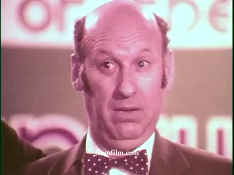 The still features a middle-aged man with a surprised expression, wearing a bow tie and a suit. The background suggests a game show setting, with colorful branding that likely relates to United Airlines. The image captures the playful and engaging tone typical of 1970s commercials, emphasizing excitement and competition.