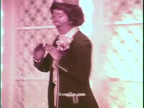 The still from the 16mm print of 'United Airlines - Deal of the Century' features a performer, likely a game show contestant or host, dressed in a vintage outfit typical of the 1970s. The background is brightly lit, possibly pink or warm-toned, creating a lively atmosphere. The individual appears animated, possibly expressing excitement or surprise, which aligns with the energetic tone of a game show commercial from that era. The overall composition suggests a playful and engaging promotional context.