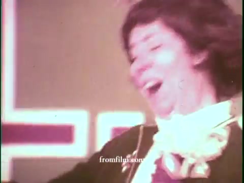 The still captures a vibrant moment from a 1970s game show commercial for United Airlines. A contestant or host appears animated and joyful, likely celebrating a win or announcement. The background features bold, colorful graphics typical of the era, contributing to the energetic atmosphere of the commercial. The overall mood suggests excitement and enthusiasm, reflective of game shows from that time period.