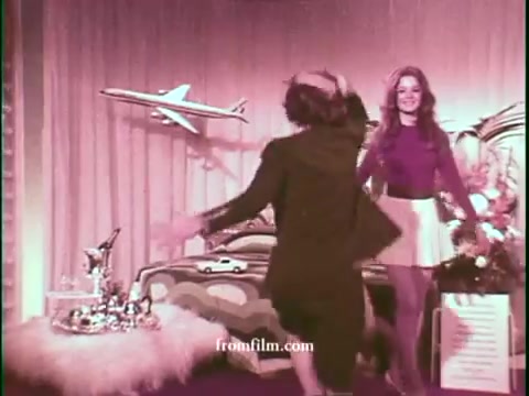 The still features a lively game show set with a colorful backdrop. A woman in a stylish outfit engages energetically with another person, suggesting a playful atmosphere. In the background, a model airplane hangs overhead, symbolizing travel. The décor includes whimsical elements typical of 1970s television, creating a vibrant and nostalgic feel.