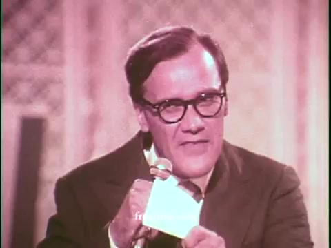 The still features a male host with glasses, dressed in a suit, holding a card and speaking into a microphone. The background suggests a game show setting, with a warm, vintage color palette typical of the 1970s. The overall tone is engaging, reflecting the promotional nature of the commercial for United Airlines.