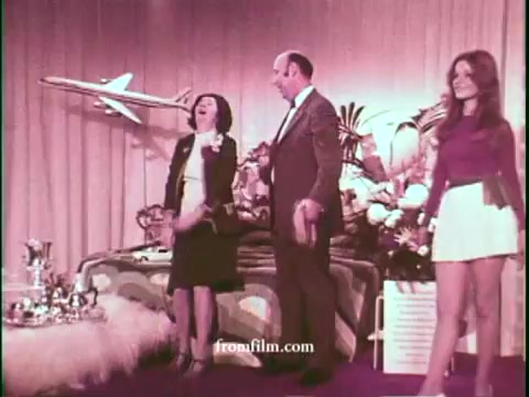 The still features a brightly colored game show set from the 1970s, showcasing a jovial host and two contestants. One woman, dressed in a stylish outfit, expresses excitement, while the man interacts with an audience member. In the background, a model airplane is prominently displayed, alongside luxurious props and floral arrangements, emphasizing the glamorous appeal of United Airlines. The overall ambiance reflects the vibrant and playful style typical of the era's television commercials.