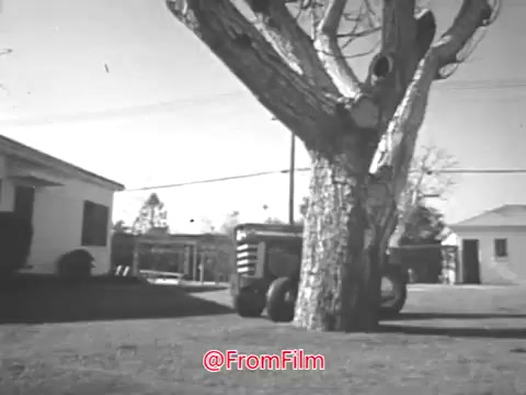 The still from the Massey Ferguson lawn tractor commercial features a vintage 16mm black-and-white scene. It shows a Massey Ferguson lawn tractor parked on a grassy area near a tree, with residential structures in the background. The setting appears to be suburban, typical of the 1960s aesthetic. The visual emphasizes the lawn tractor's practicality for homeowners, aligning with the commercial’s promotional intent. The overall tone is nostalgic and highlights outdoor domestic life.   For licensing and higher resolution options, you can contact the source for details.