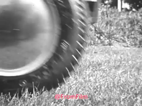 The still captures a close-up view of a tire of a Massey Ferguson lawn tractor as it rolls over grass. The emphasis is on the tire's tread, showcasing its design and interaction with the lawn. The black and white aesthetic reflects the 1960s style, evoking a nostalgic feel associated with vintage commercials. The context suggests this is part of a promotional effort featuring Jerry Colonna, likely highlighting the efficiency or performance of the lawn tractor.