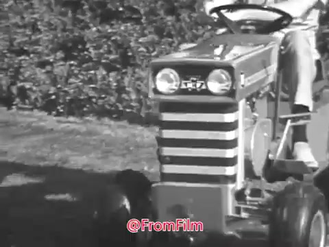 The still depicts a Massey Ferguson lawn tractor being driven, showcasing its distinctive design from the 1960s. The tractor features simple lines and a unique striped pattern on its front, indicating the brand's style. In the background, there are neatly trimmed hedges, suggesting a well-maintained lawn setting. The overall aesthetic captures a nostalgic look at gardening and machinery from that era.