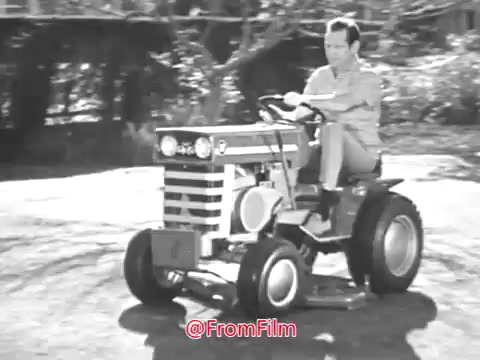 The still depicts a vintage Massey Ferguson lawn tractor being driven by Jerry Colonna, set in a sunny outdoor environment. Colonna is seen seated on the tractor, which features a distinctive design typical of 1960s machinery, characterized by its robust build and prominent front grille. The scene likely highlights the ease and enjoyment of using the tractor for lawn care, aligning with the commercial's promotional message. The image is in black and white, capturing the nostalgic essence of that era.