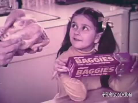 The still from the 'Baggies alligator storage bags' commercial features a young girl holding a box of Baggies alligator bags while a man presents a chicken. The setting appears to be a kitchen, emphasizing the practical use of the storage bags for food. The girl's expression suggests curiosity or excitement, effectively conveying the commercial's focus on freshness and protection against freezer burn. The alligator theme adds a playful element to the product's rugged characteristics.