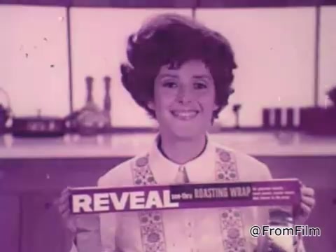 In this still from the 'Baggies alligator storage bags' commercial, a woman with a bright smile holds up a box of 'REVEAL low-dry roasting wrap' against a kitchen backdrop. Her hairstyle is styled in a retro fashion, contributing to the commercial's vintage aesthetic. The overall atmosphere suggests a focus on home cooking and food preservation, emphasizing the reliability of the product for keeping food fresh.