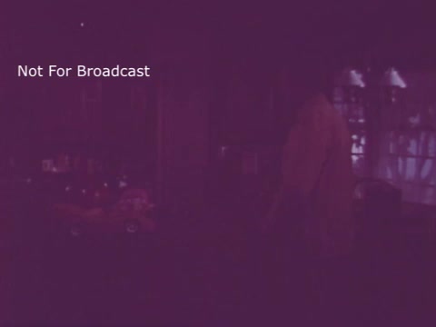 The still from the 16mm print of 'Toy Safety - Consumer Product Safety Commission PSA' from the 1970s features a dimly lit interior, possibly a living room or play area. A figure is seen standing, possibly a parent or guardian, who appears to be looking at a collection of toys. The overall atmosphere suggests a focus on the importance of toy safety in a domestic setting, highlighting vigilance for hazardous playthings. The watermark indicates that the image is not intended for broadcast.